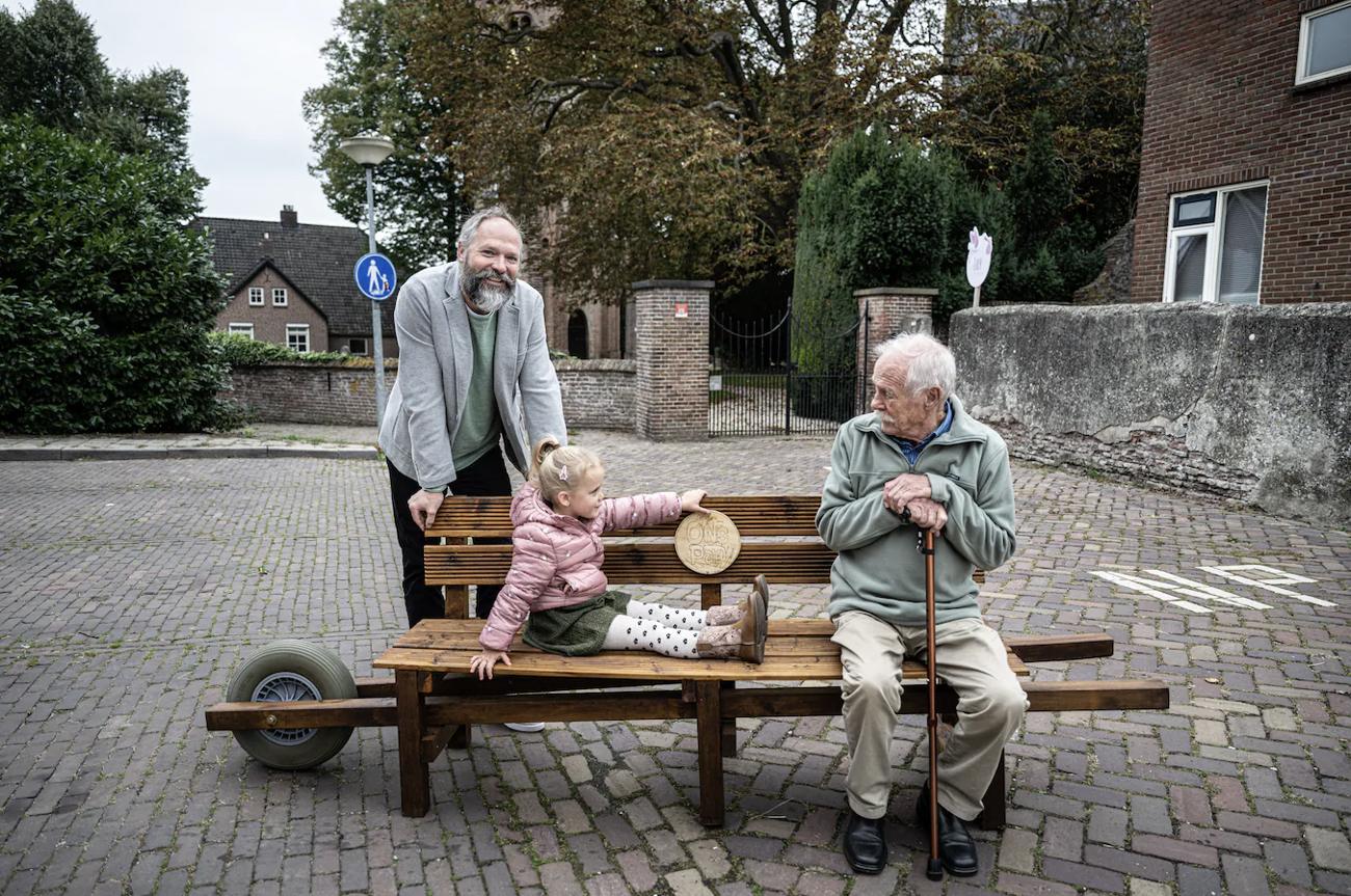Ivo Wouters staat achter de rollende buurtbank in de straat, zijn dochter zit op de bank en een oudere buurman met wandelstok zit naast haar